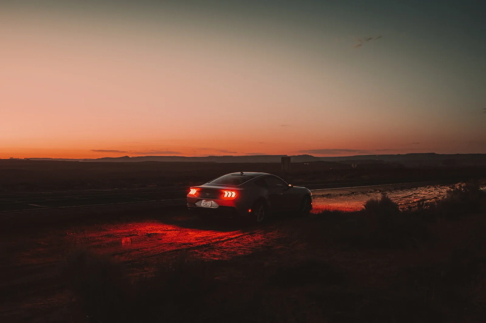 A car with glowing red taillights on a desert track beneath an orange sunset